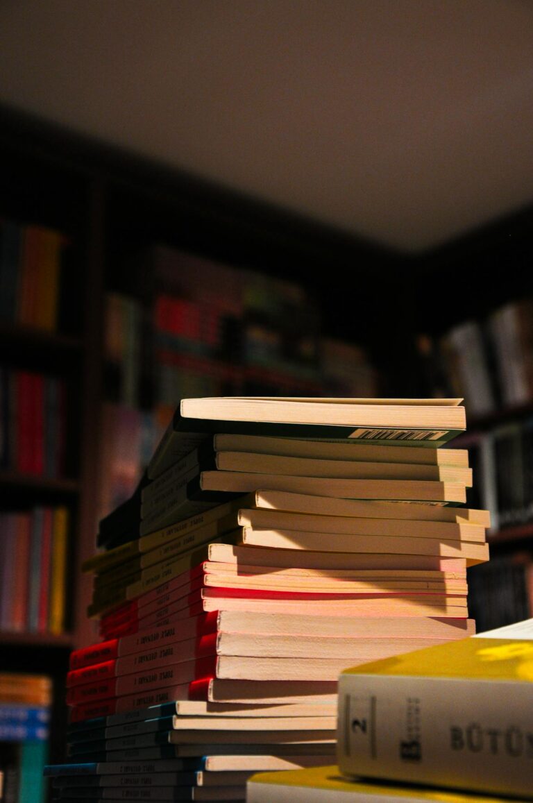 A tall stack of books in a cozy library setting with soft lighting and filled bookshelves in the background.